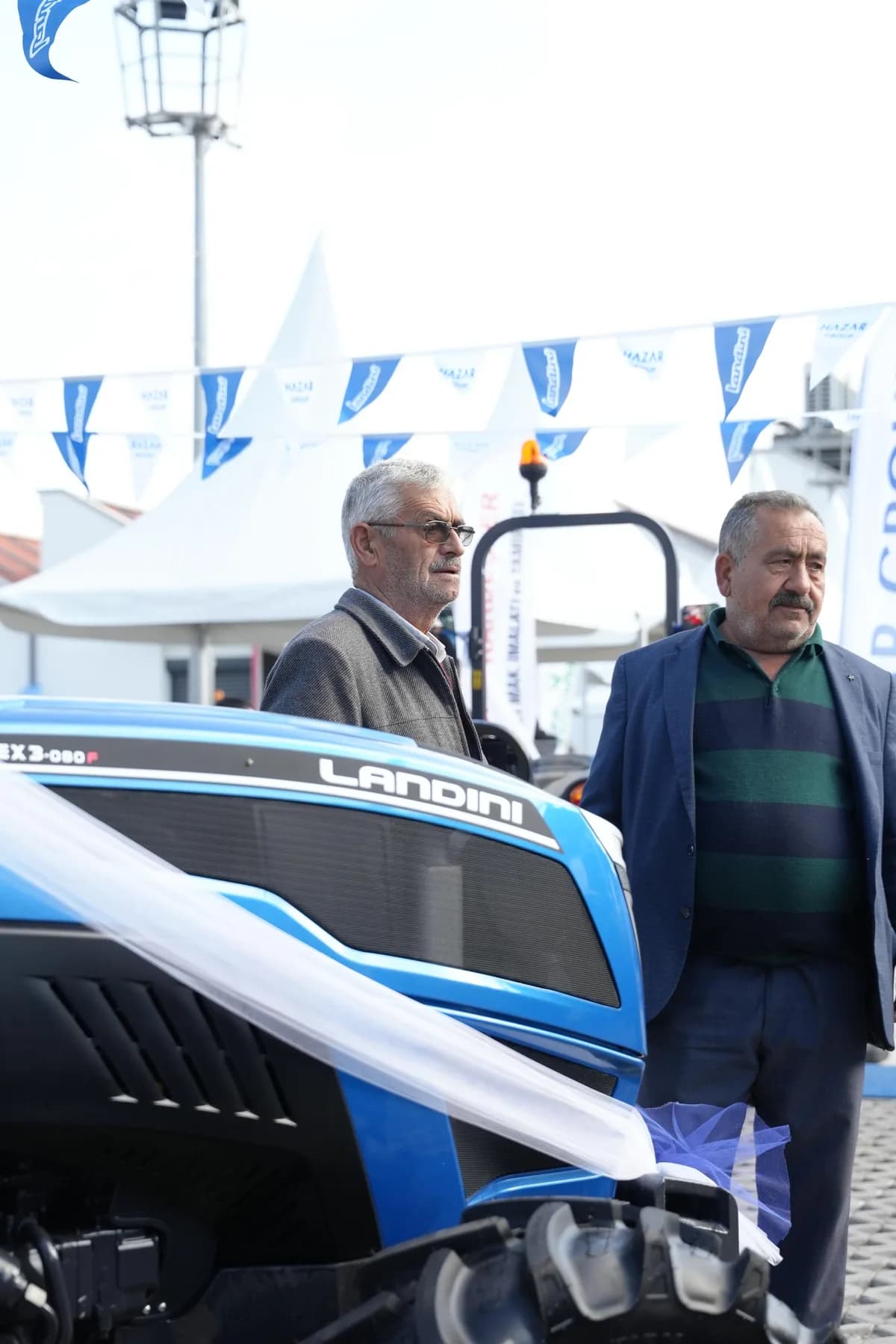 Mavi Landini traktörün önünde duran iki ziyaretçi, teslim kurdelesiyle süslenmiş makine