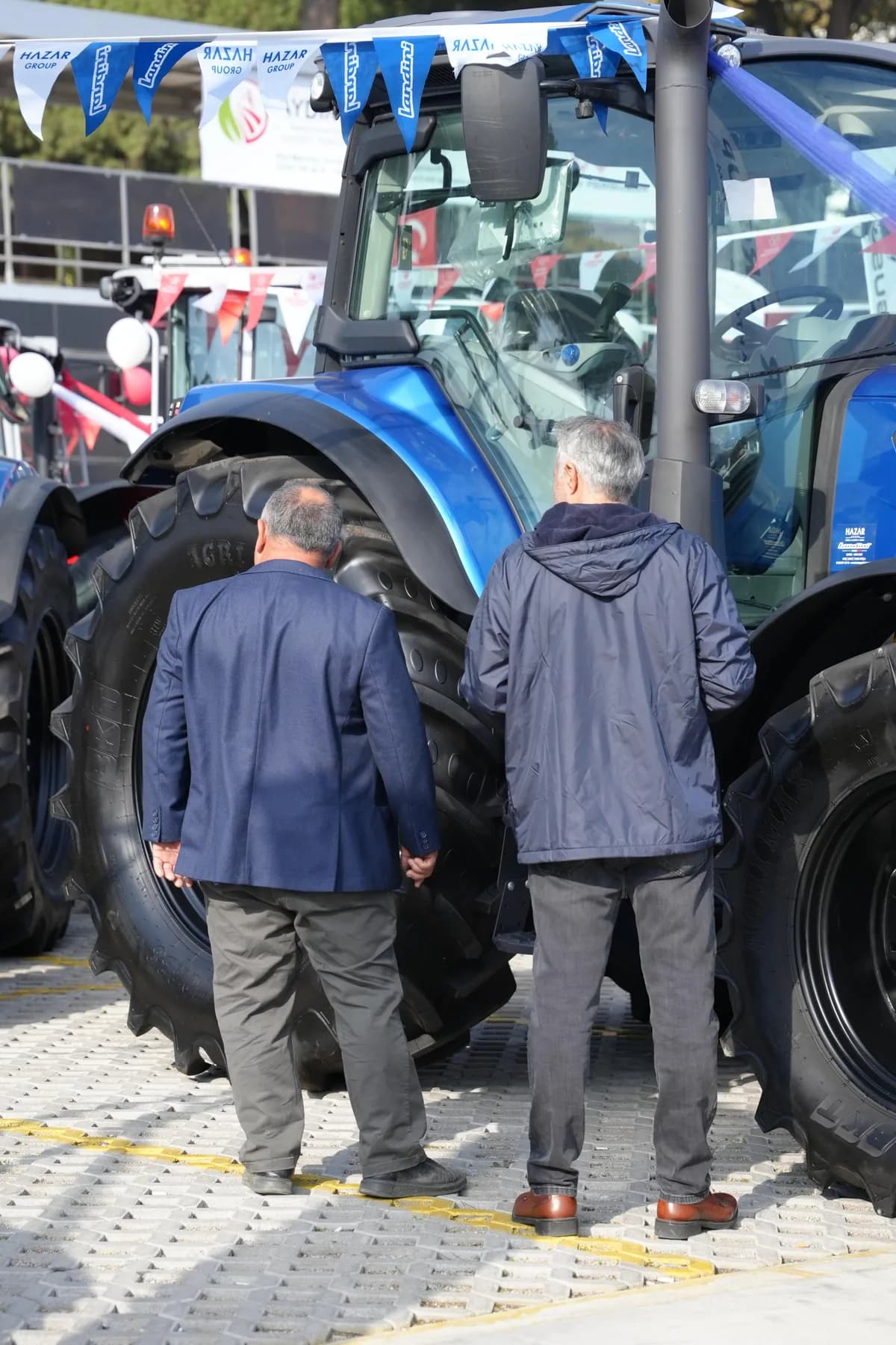 Mavi traktörü inceleyen iki kişi, Hazar Group flamalarıyla süslenmiş açık hava standı