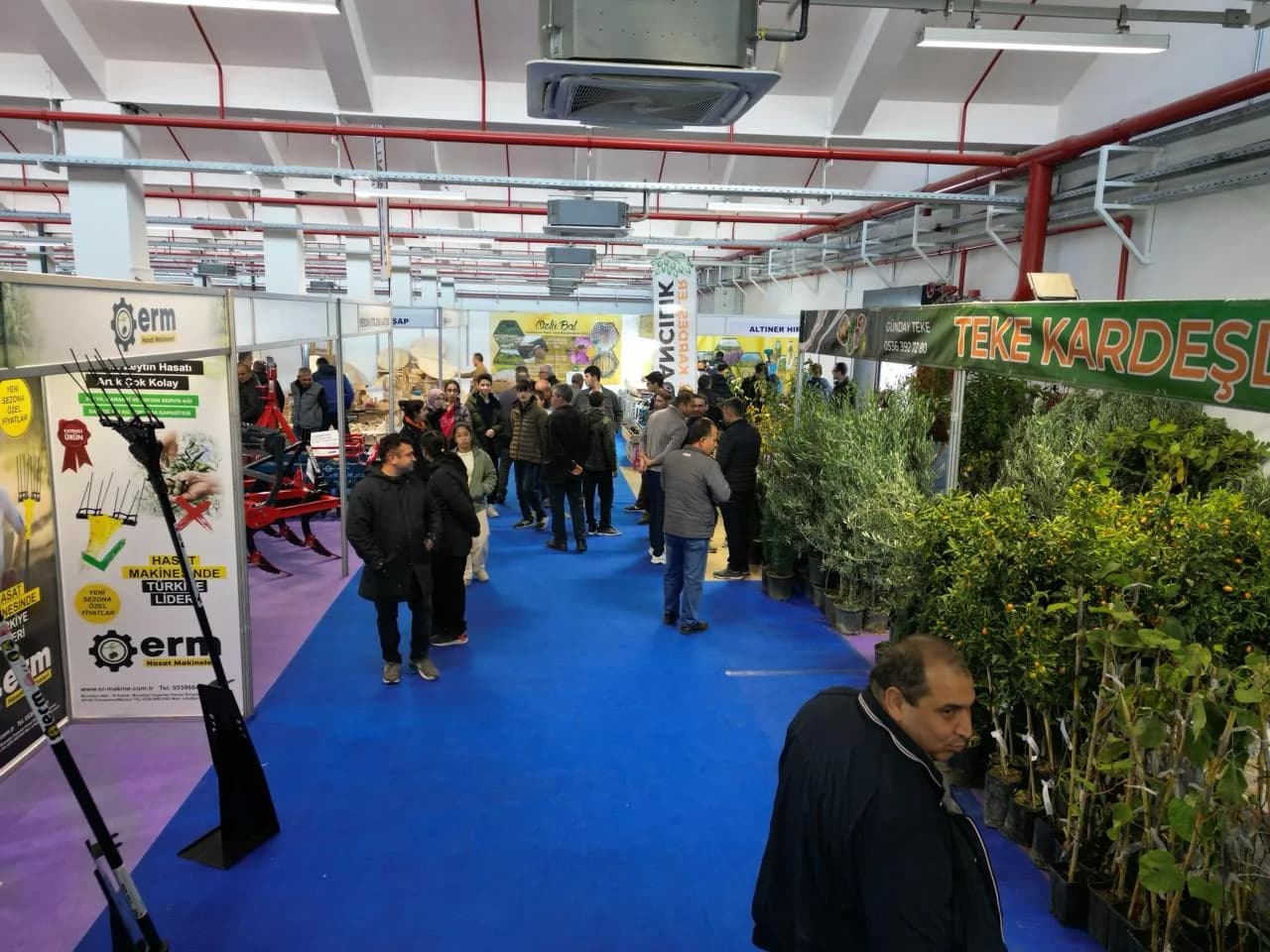 Fuar salonunda kalabalık ziyaretçi, fidan standı ve hasat makineleri afişiyle geniş koridor görünümü